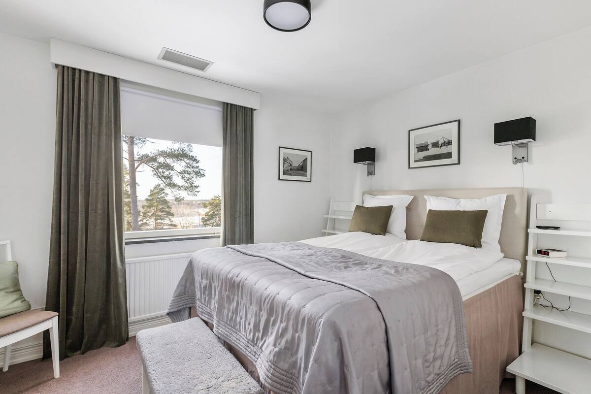 A bedroom with a large bed, a window with curtains, a bedside table, and a wall-mounted shelf.