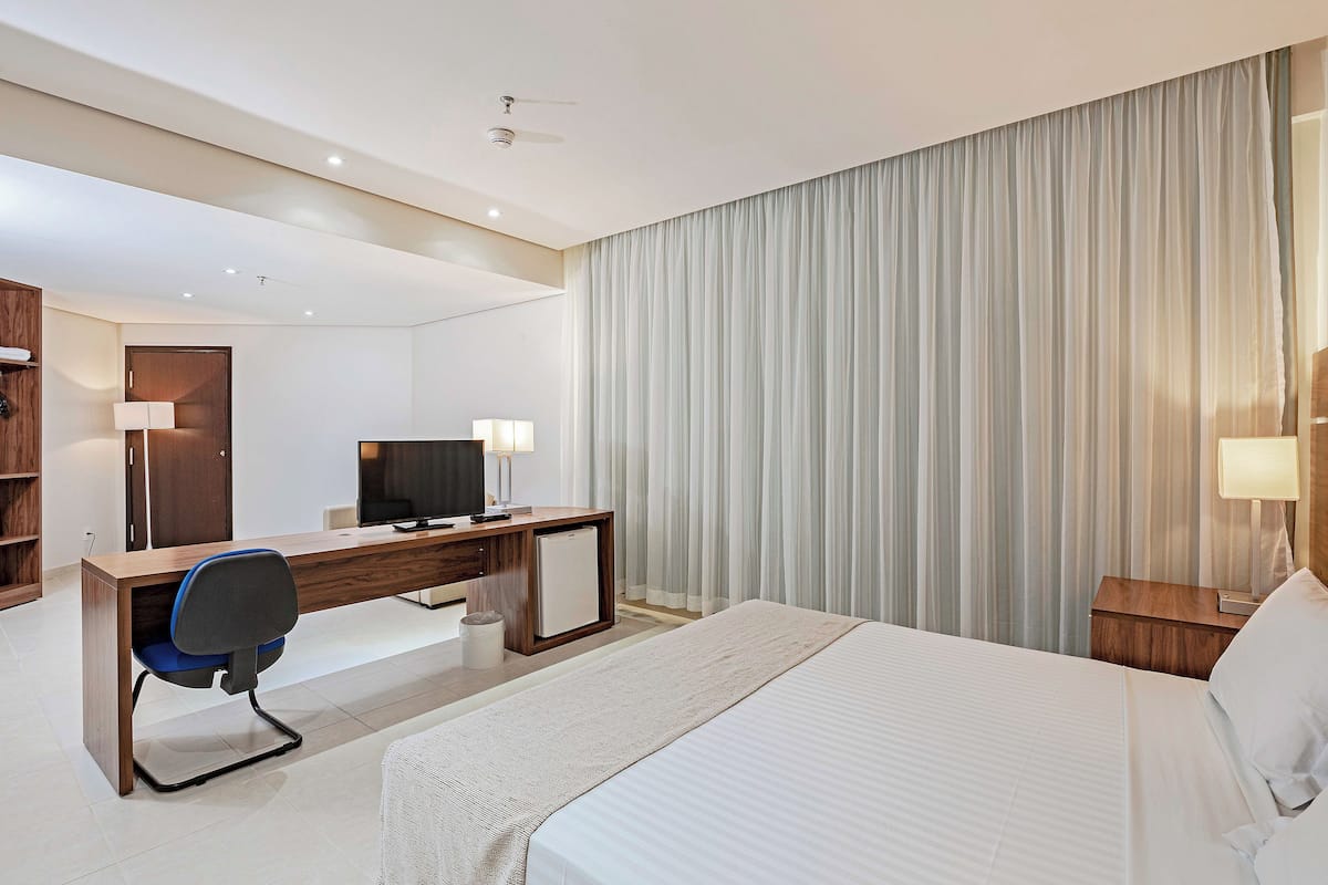 A modern hotel room with a large bed, a desk with a computer, a television, and a wardrobe.