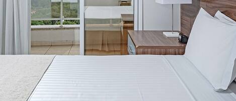 A hotel room with a bed, a nightstand with a lamp, a sliding glass door to a balcony, and a view of the cityscape.
