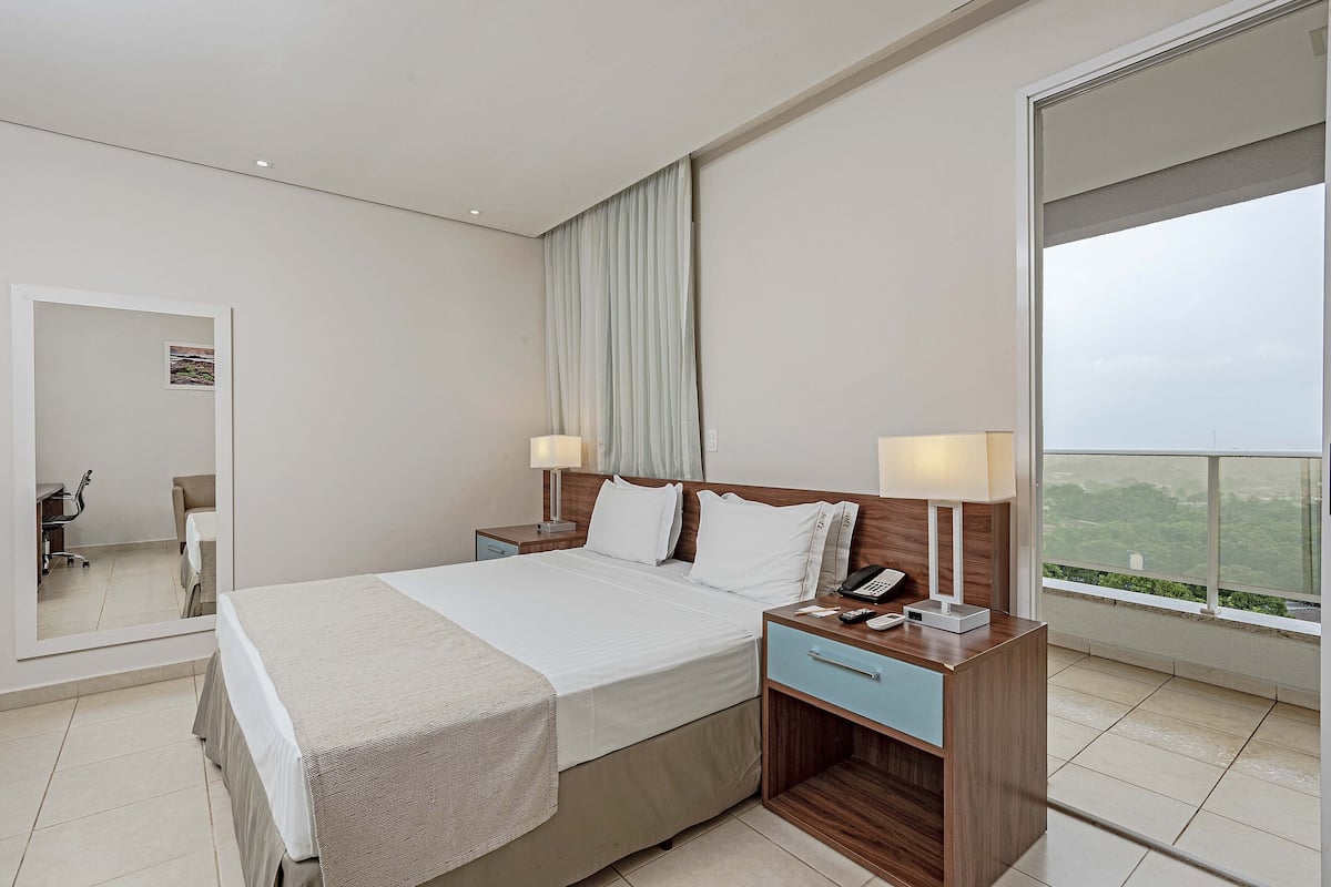 A modern hotel room with a large bed, bedside tables, a desk, and a view of the outdoors.