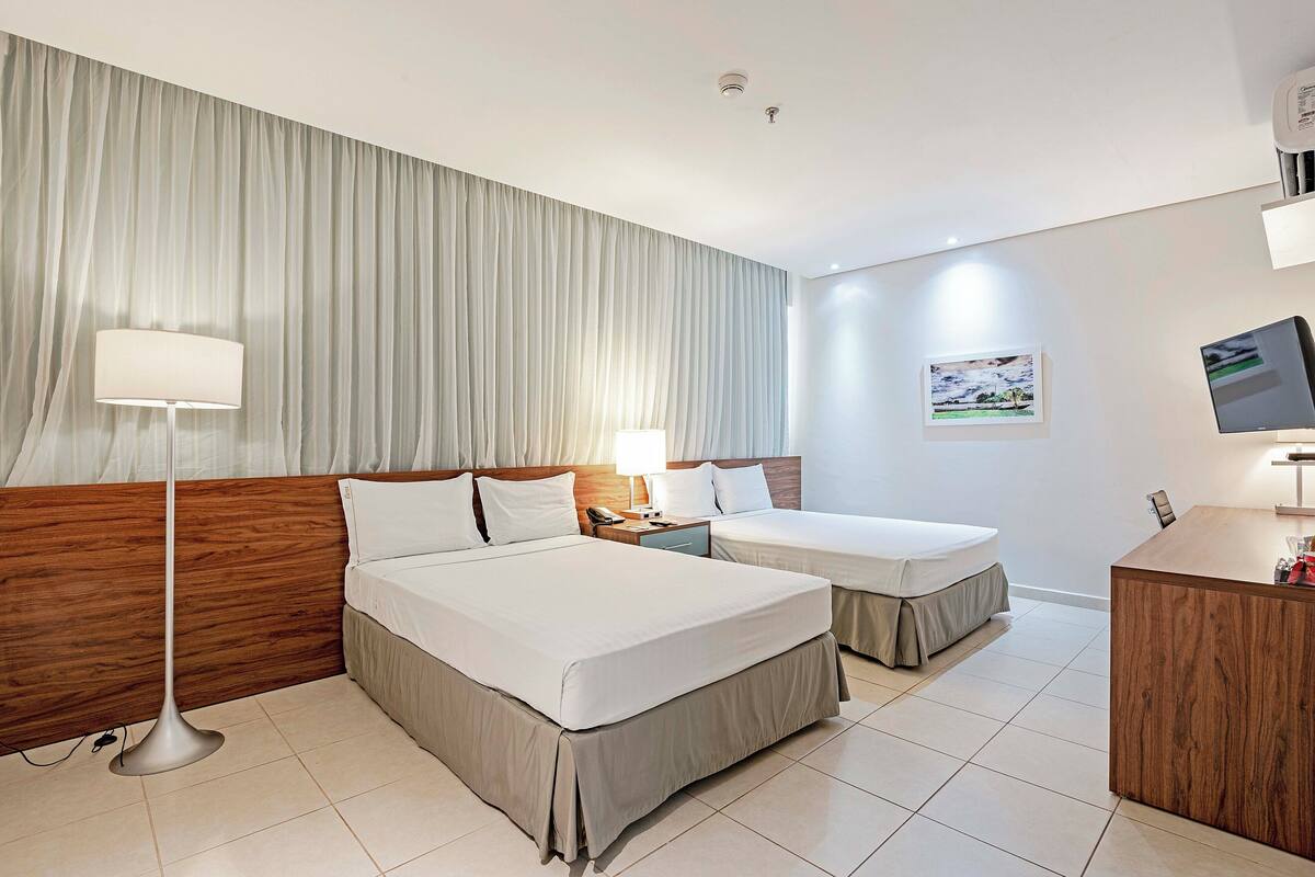 A hotel room with a large bed, a smaller bed, a desk, a lamp, a phone, and a picture on the wall.