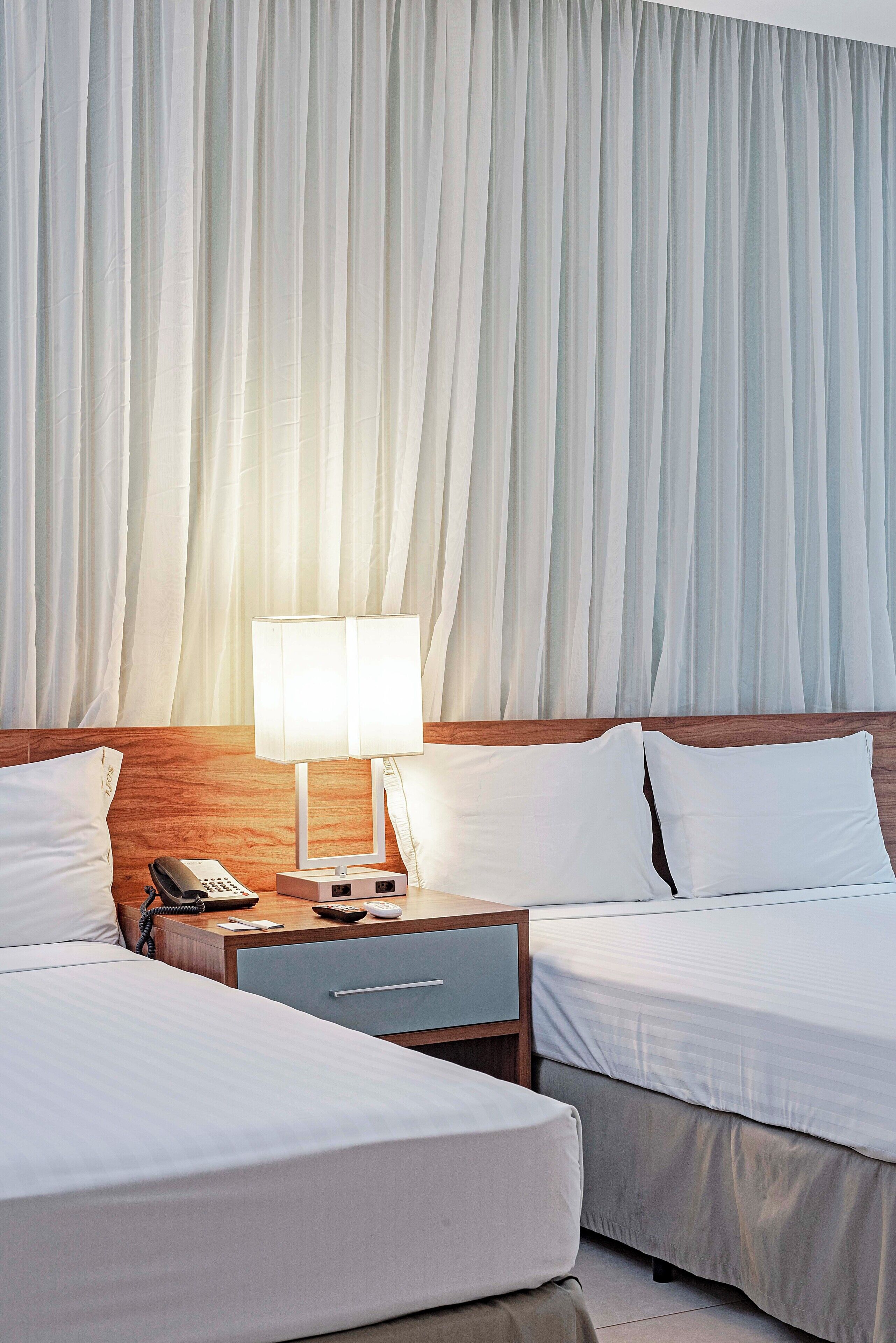A hotel room with two beds, a nightstand with a lamp, a phone, and a small drawer.