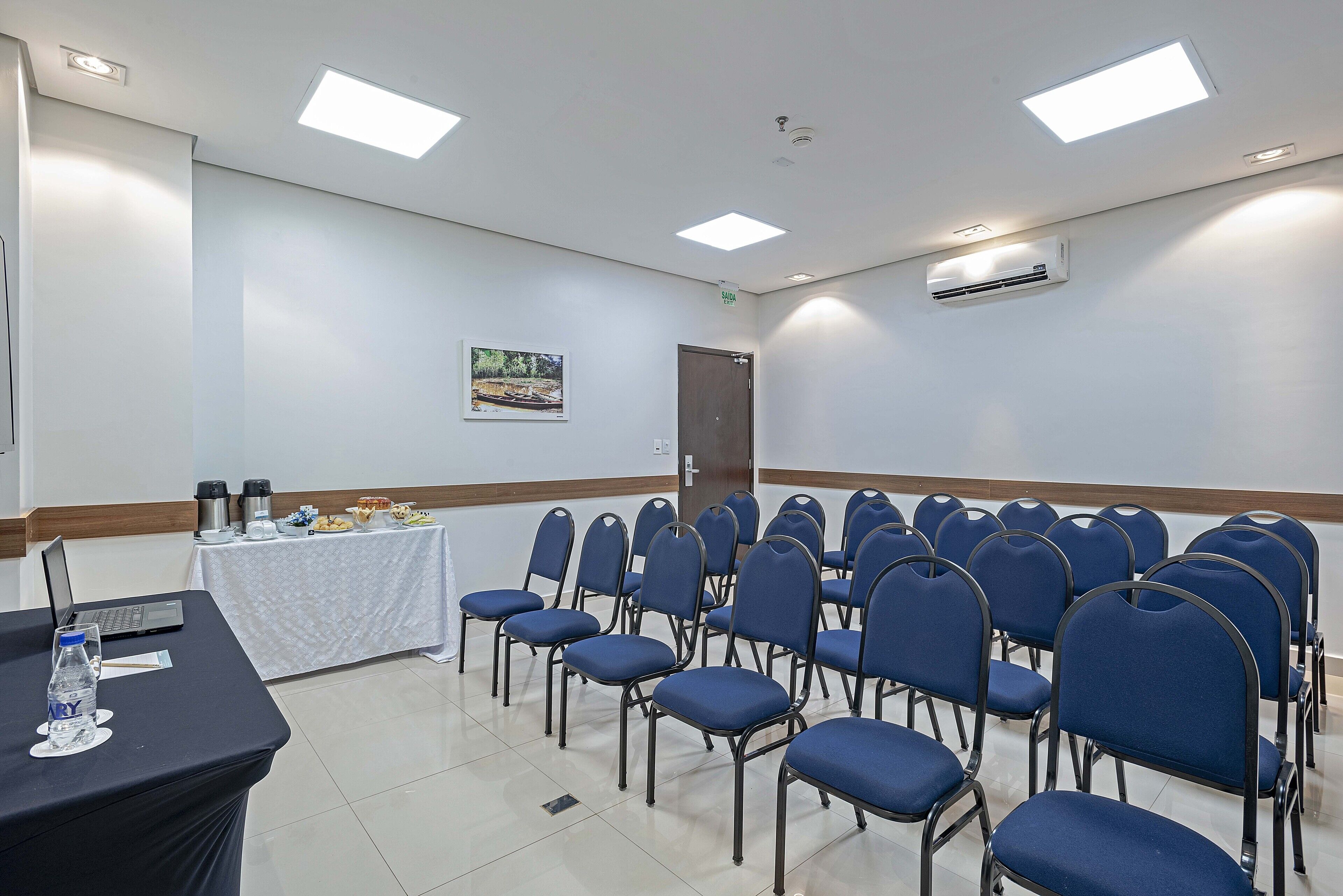 Un salón de conferencias con filas de sillas azules, una mesa con una computadora portátil y una botella de agua, y un equipo de aire acondicionado en la pared.