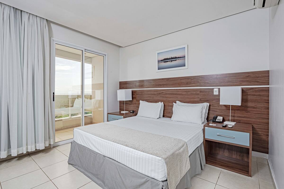 A hotel room with a large bed, a wooden headboard, a nightstand with a lamp, and a view of the outdoors through a sliding door.