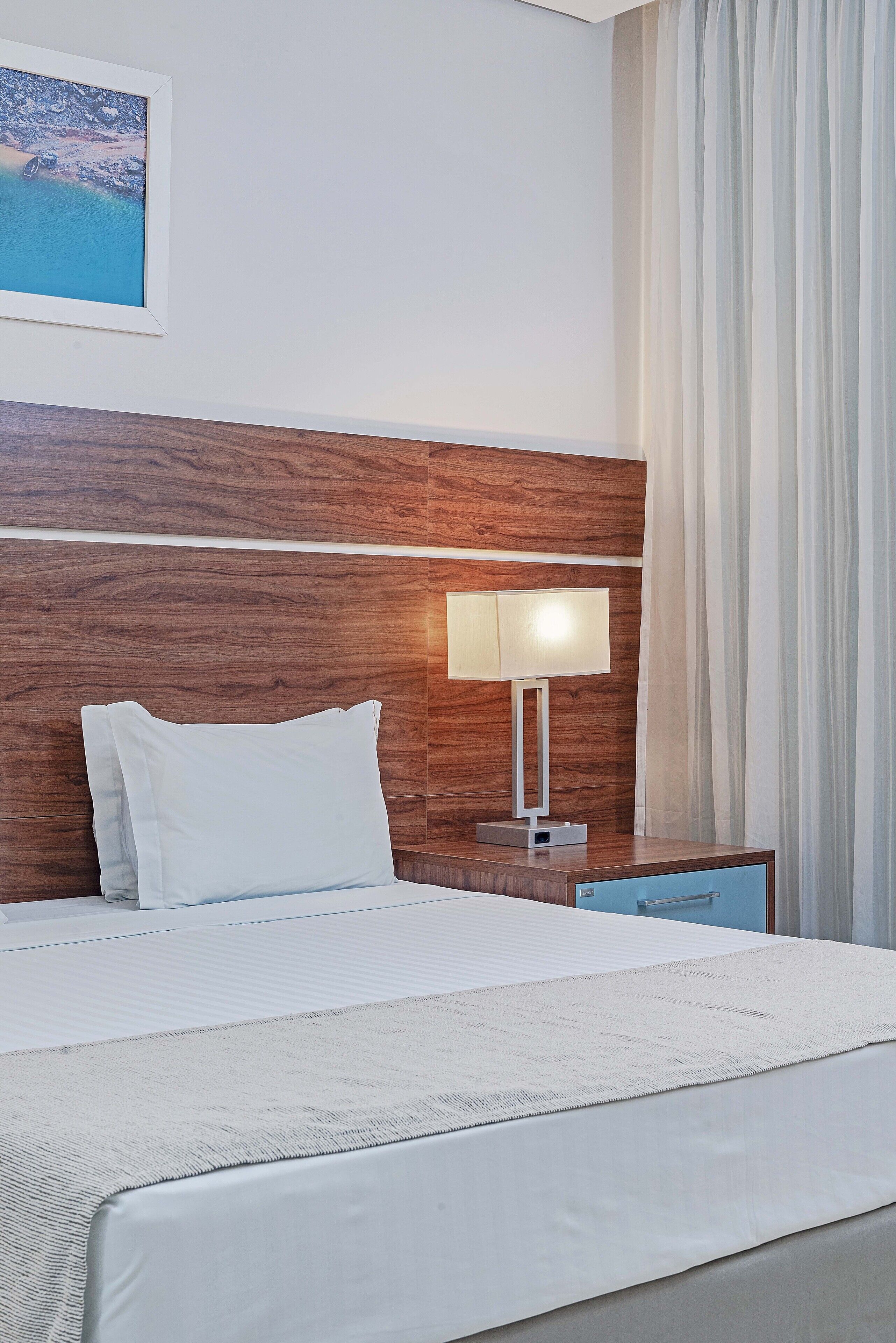 A hotel room with a wooden headboard, a bed with white bedding, a nightstand with a lamp, and a view of the sea through the window.