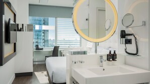 Habitación de hotel moderna con una cama grande, un espejo redondo, un lavabo blanco y vistas de la ciudad a través de amplios ventanales.