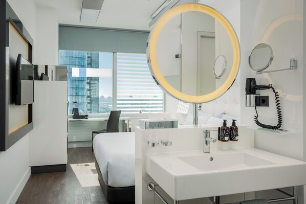 Ein modernes Hotelzimmer mit einem großen Bett, einem runden Spiegel, einem weißen Waschbecken und Blick auf die Stadt durch große Fenster.
