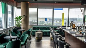 Zona de bar moderna con asientos verdes, barra y amplios ventanales con vistas a la ciudad.