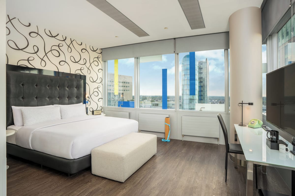 A modern hotel room with a large bed, a glass dining table, and a city view.