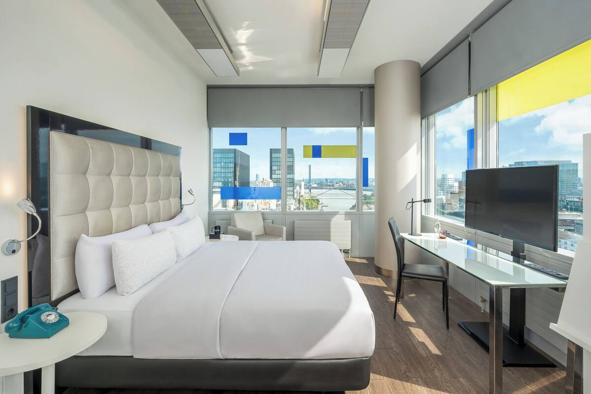 A modern hotel room with a large bed, a desk, a TV, and a city view.