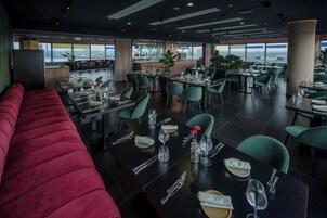Un ristorante moderno con tavoli e sedie alti, una panca imbottita rossa e vista sulla città attraverso ampie finestre.