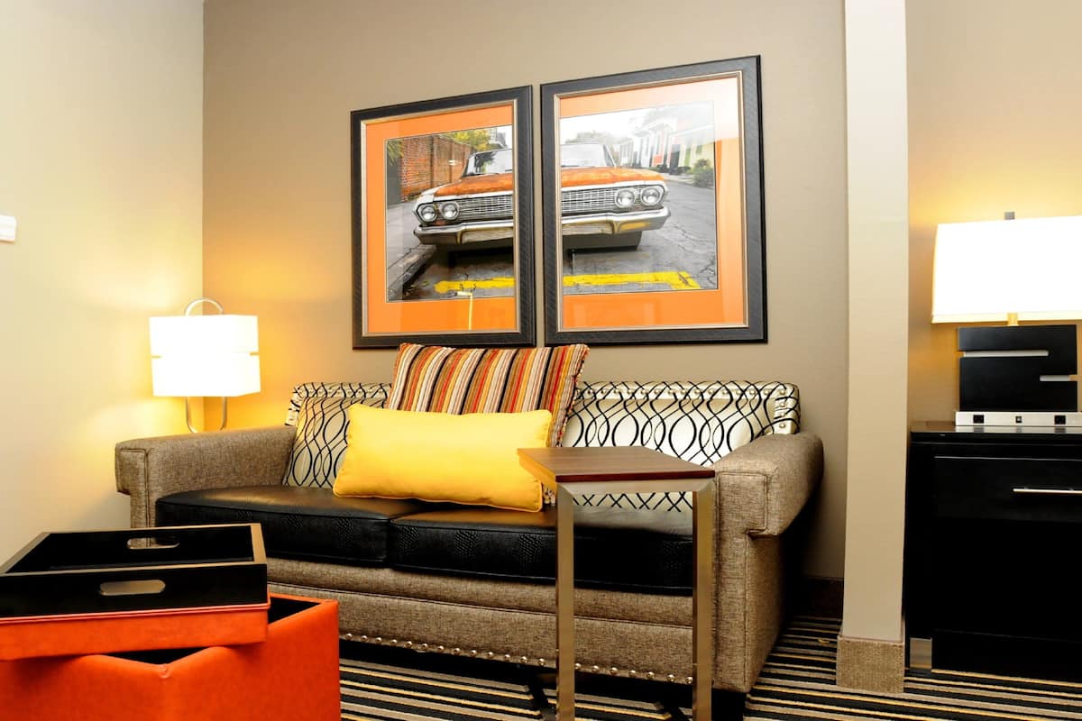 A hotel room with a sofa, a small table, a lamp, a clock, and two framed pictures on the wall.