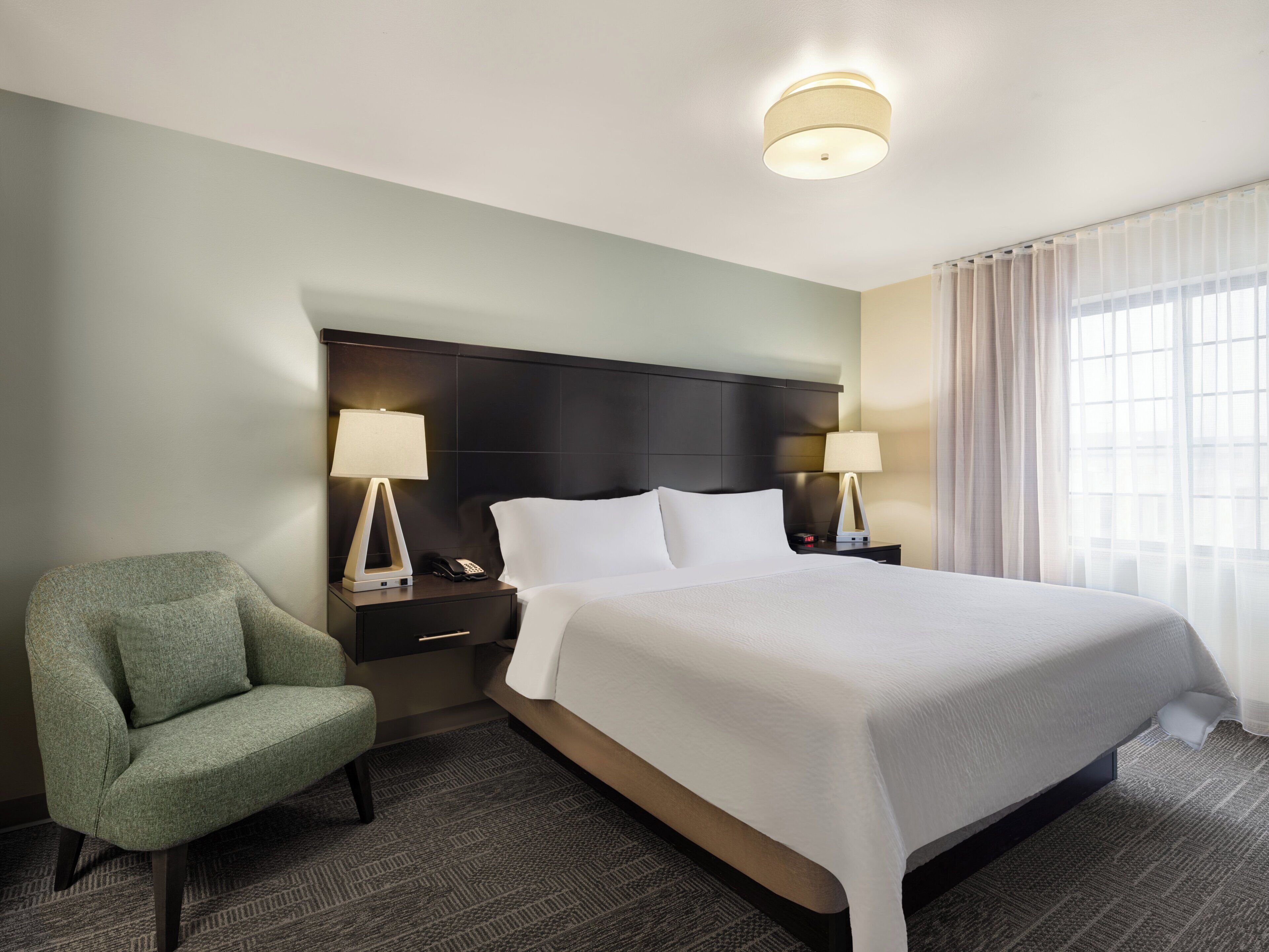 Una habitación de hotel con una cama, dos mesitas de noche, una silla y una lámpara en el techo.