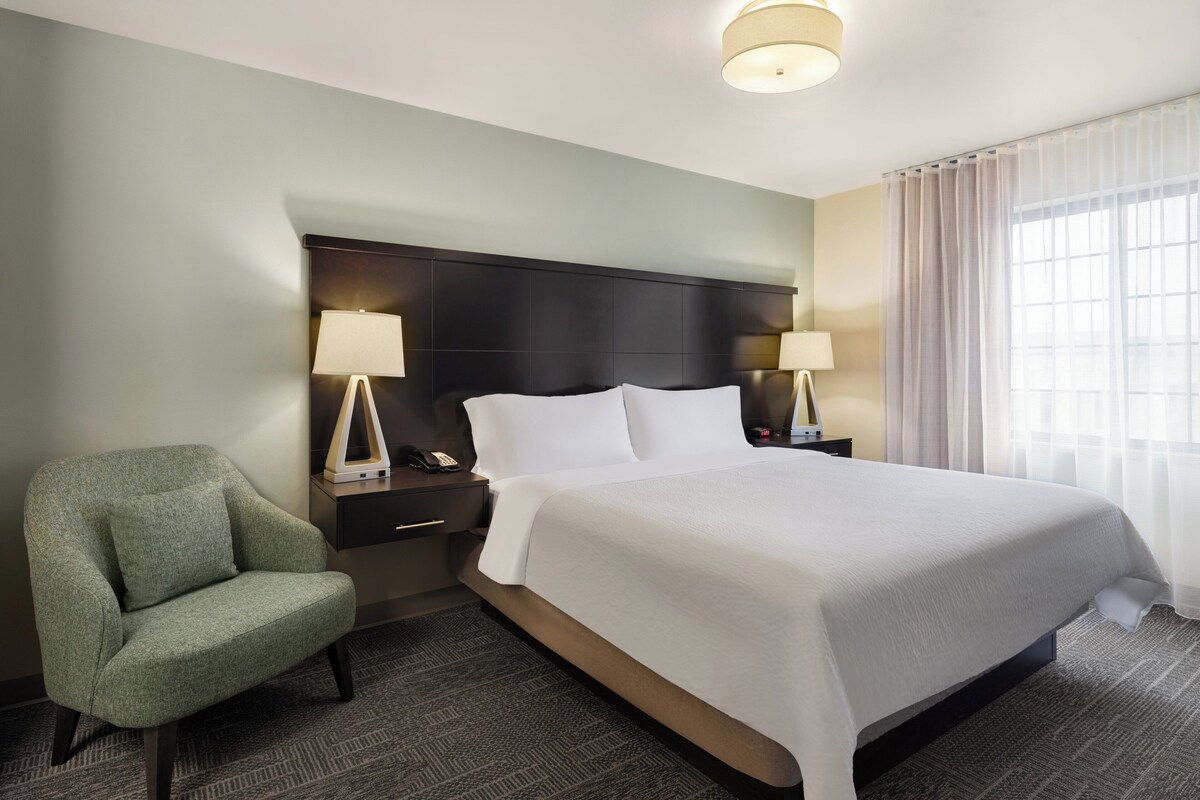 Una habitación de hotel con una cama, dos mesitas de noche, una silla y una lámpara en el techo.