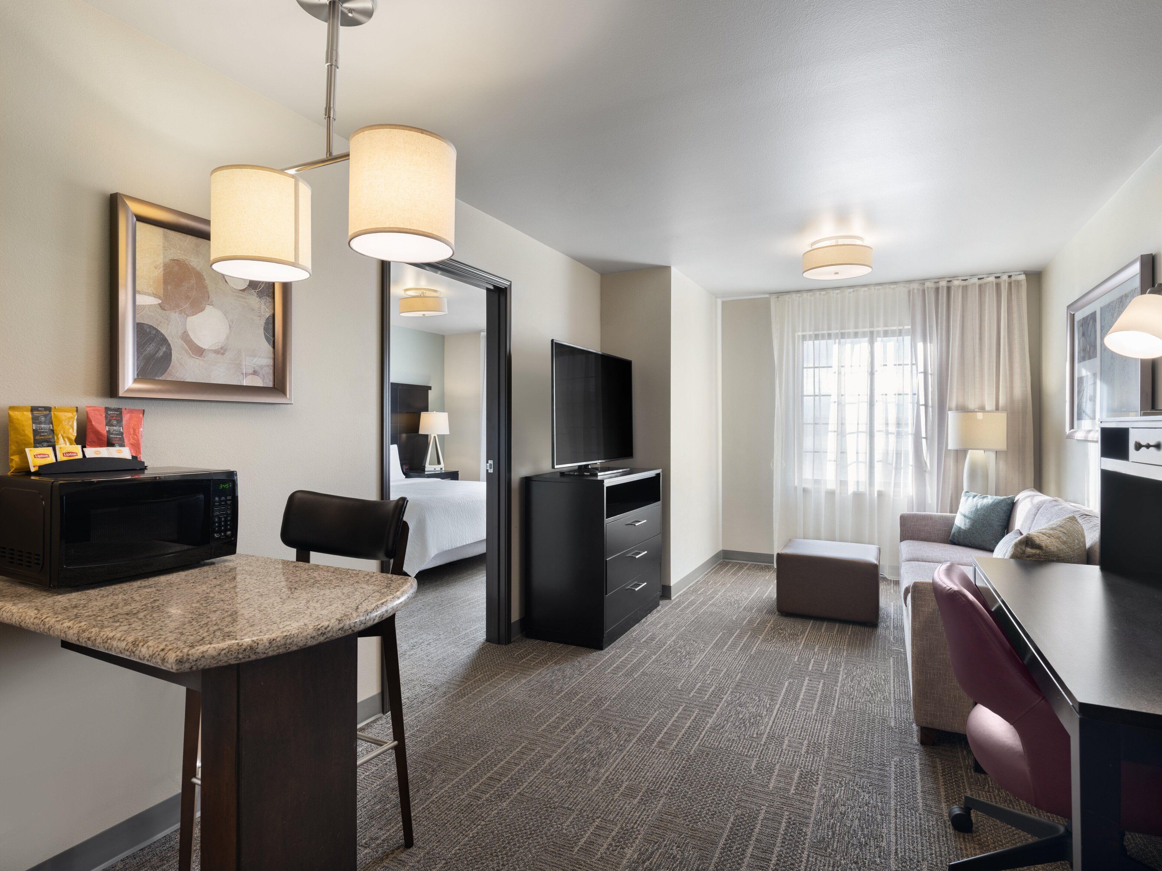 Hotelkamer met kitchenette, flatscreen televisie, een bank, bureau en groot raam met gordijnen.