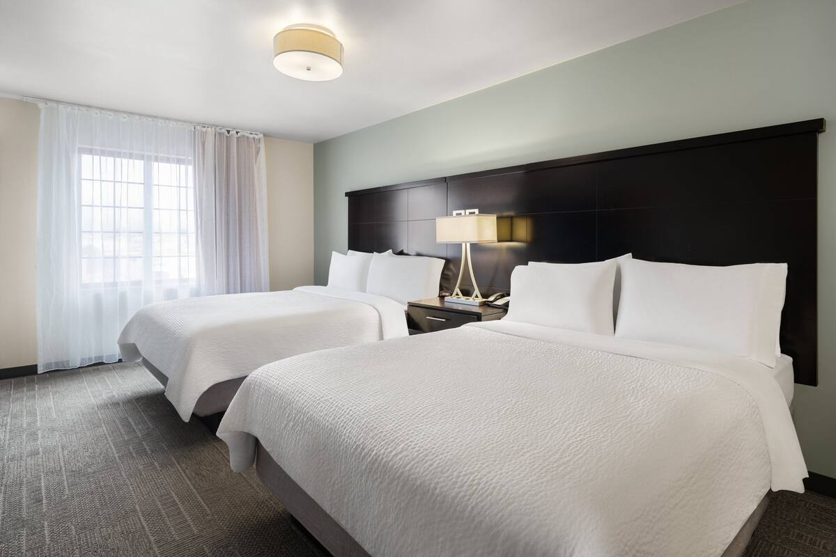 A hotel room with two beds, a nightstand with a lamp, and a large window with sheer curtains.