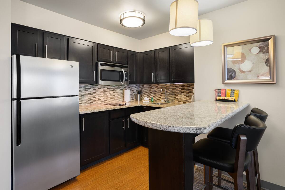 A modern kitchen with dark cabinets, stainless steel appliances, a granite countertop, and a bar area with stools.