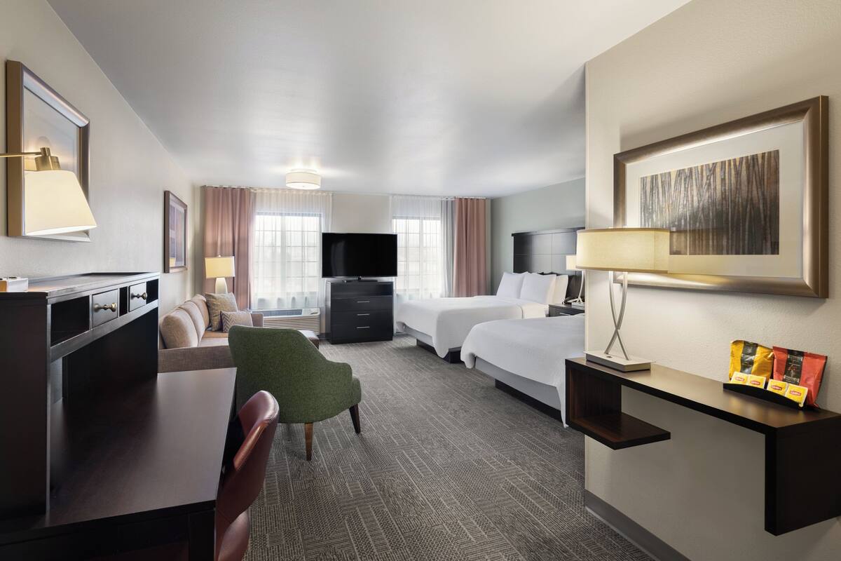 A hotel room with a desk, two beds, a TV, a green chair, and a framed picture on the wall.