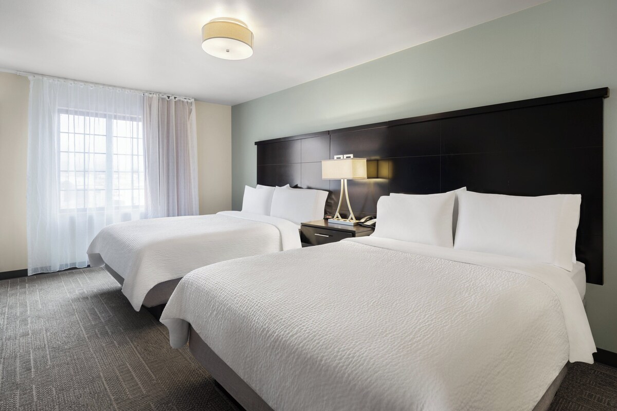 Habitación de hotel con dos camas, una mesita de noche con lámpara y un ventanal con cortinas translúcidas.