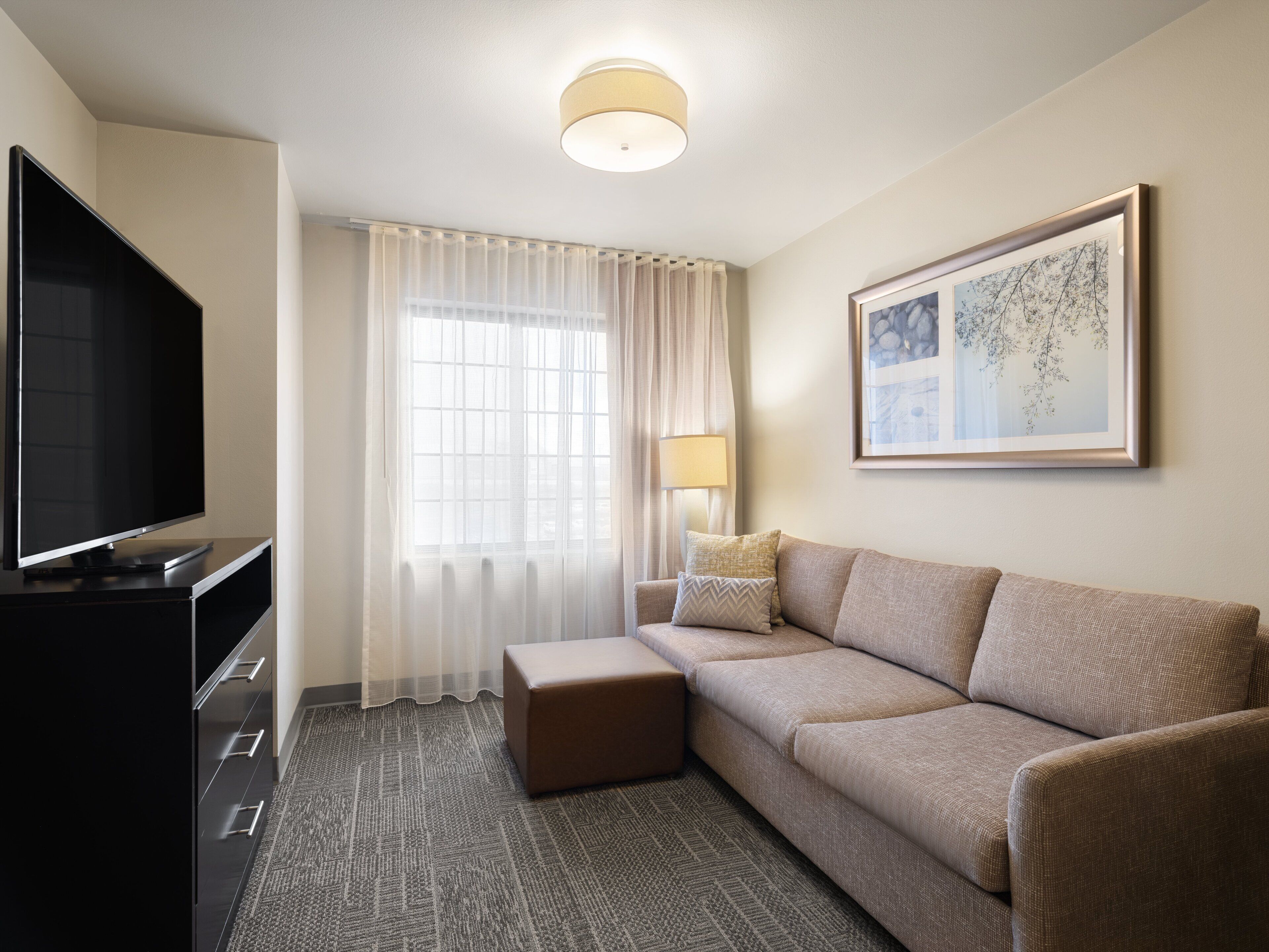 Una camera d'albergo moderna con televisione a schermo piatto, un divano, un tavolino con una lampada e un quadro incorniciato appeso al muro.