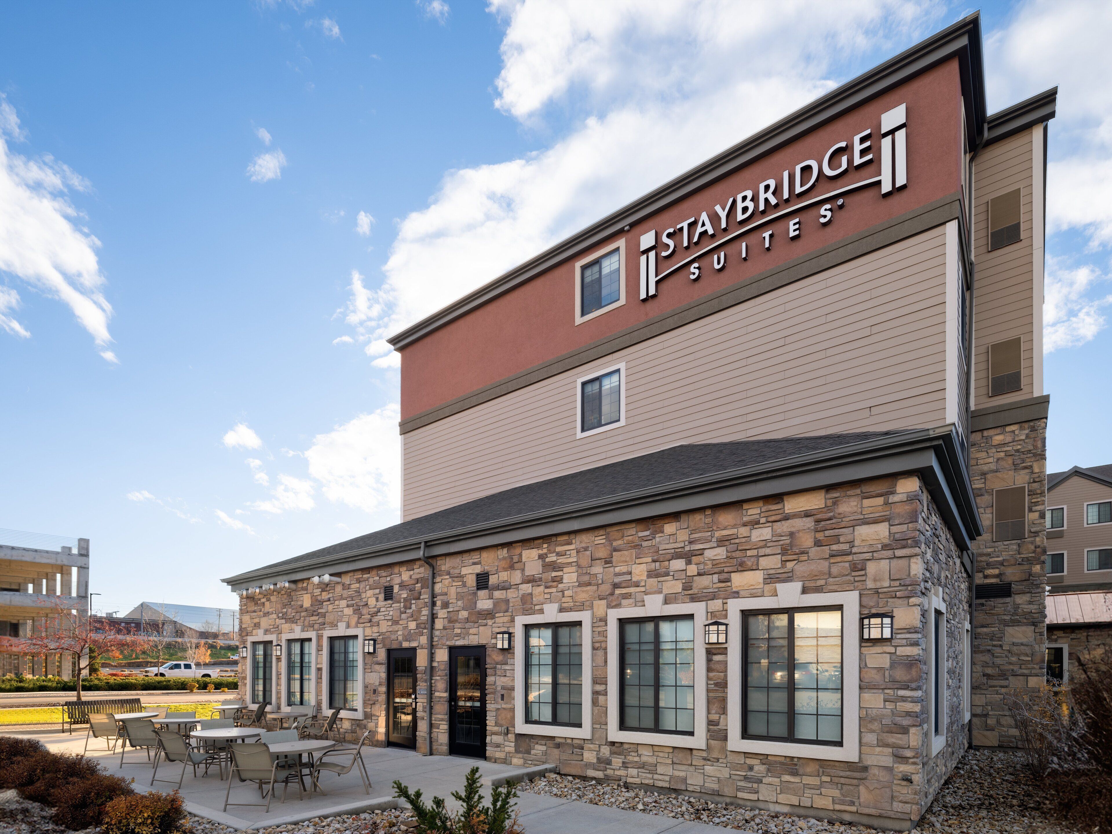 A Staybridge Suites hotel with a stone facade, outdoor seating area, and a clear sky.