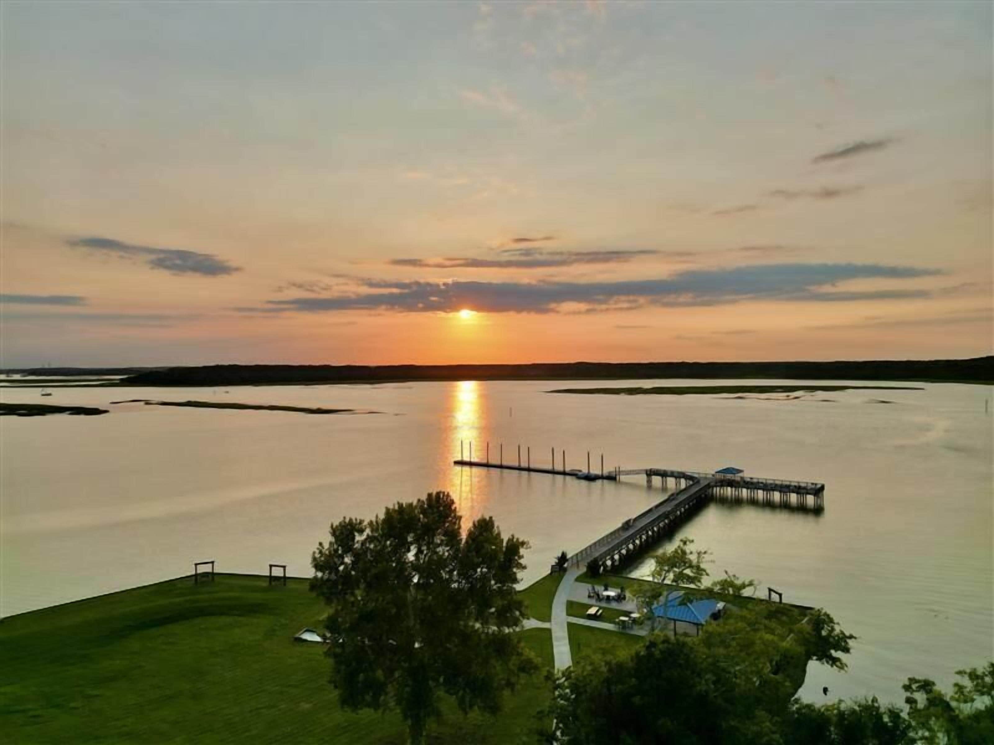 A sunset over a calm lake with a pier extending into the water, surrounded by grassy areas and trees.