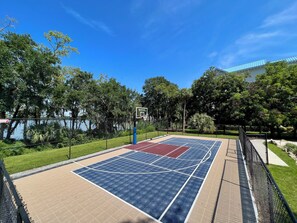 An outdoor basketball court with a chain-link fence, surrounded by trees and a building in the background.