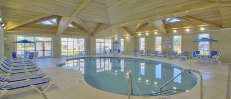 Una piscina interior con sillas de descanso azules y blancas, una escalera de piscina y grandes ventanales que dejan entrar luz natural.