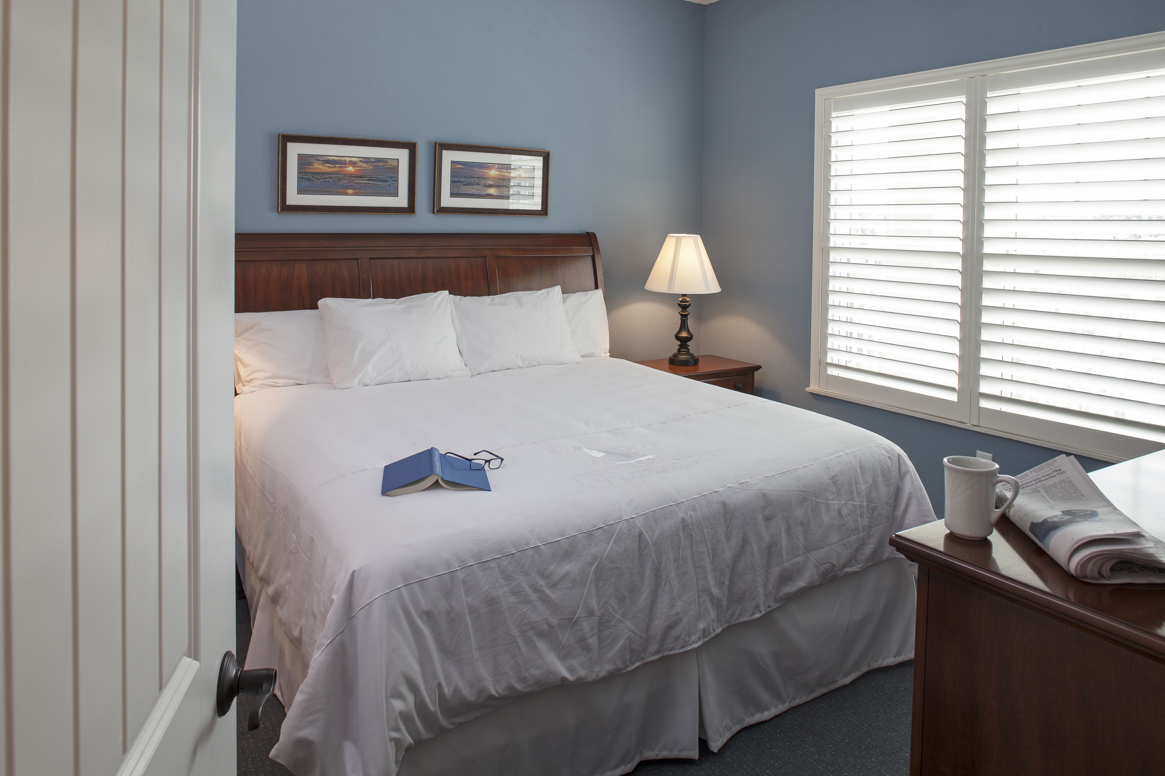 A neatly made bed with white linens, a bedside lamp, and a window with blinds.