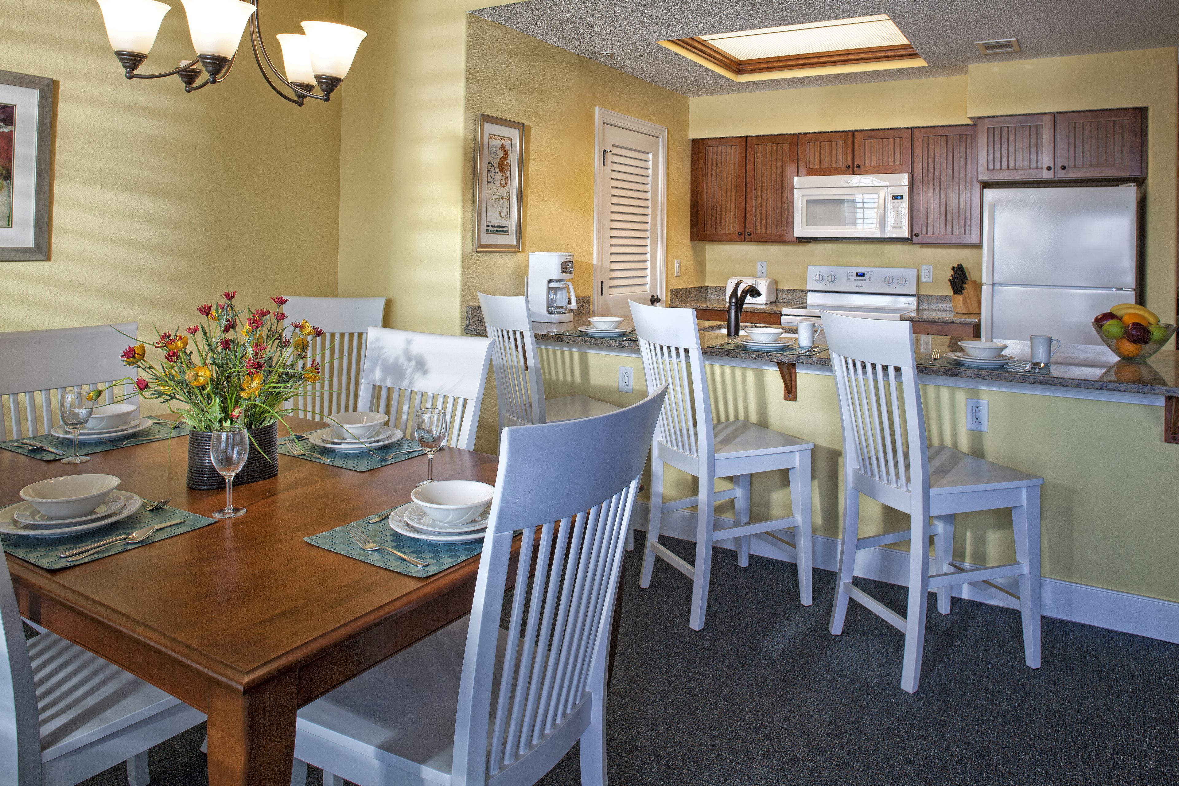Un comedor con una mesa de madera preparada para cuatro personas, sillas blancas y un jarrón de flores.