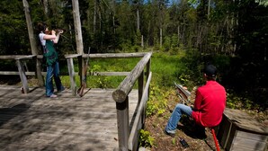 Une terrasse en bois où une personne vise avec un arc, une autre personne est assise sur un tabouret, et une zone boisée se trouve à l’arrière-plan.