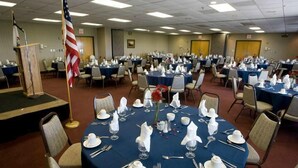 Une salle de conférence avec plusieurs tables rondes disposées pour un repas, chacune recouverte d’une nappe blanche et agrémentée d’assiettes, de couverts et de verres. Un drapeau américain est bien visible au premier plan.