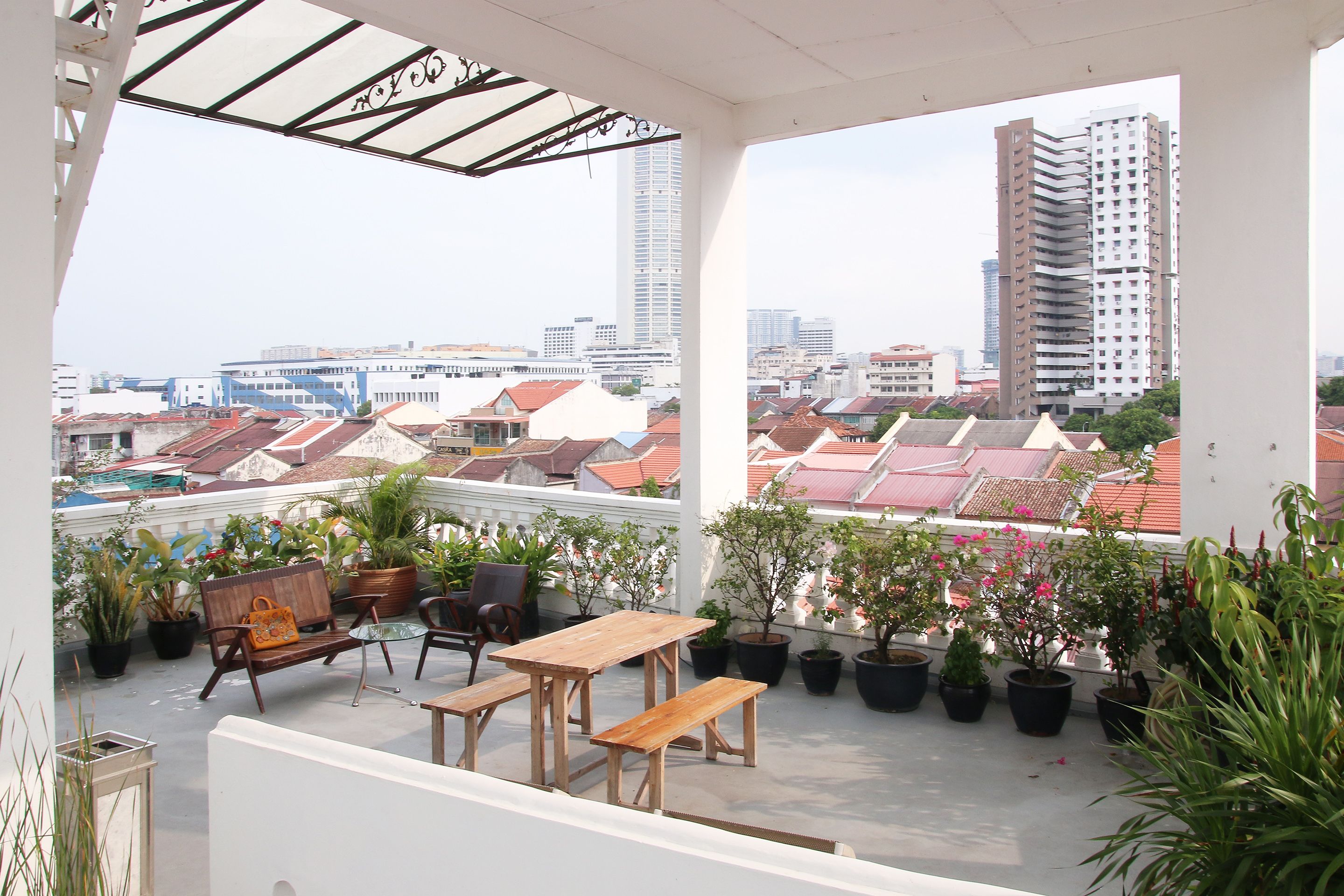 Terraza en la azotea con mesa y bancos de madera, rodeada de plantas en macetas y vistas a un paisaje urbano con edificios.