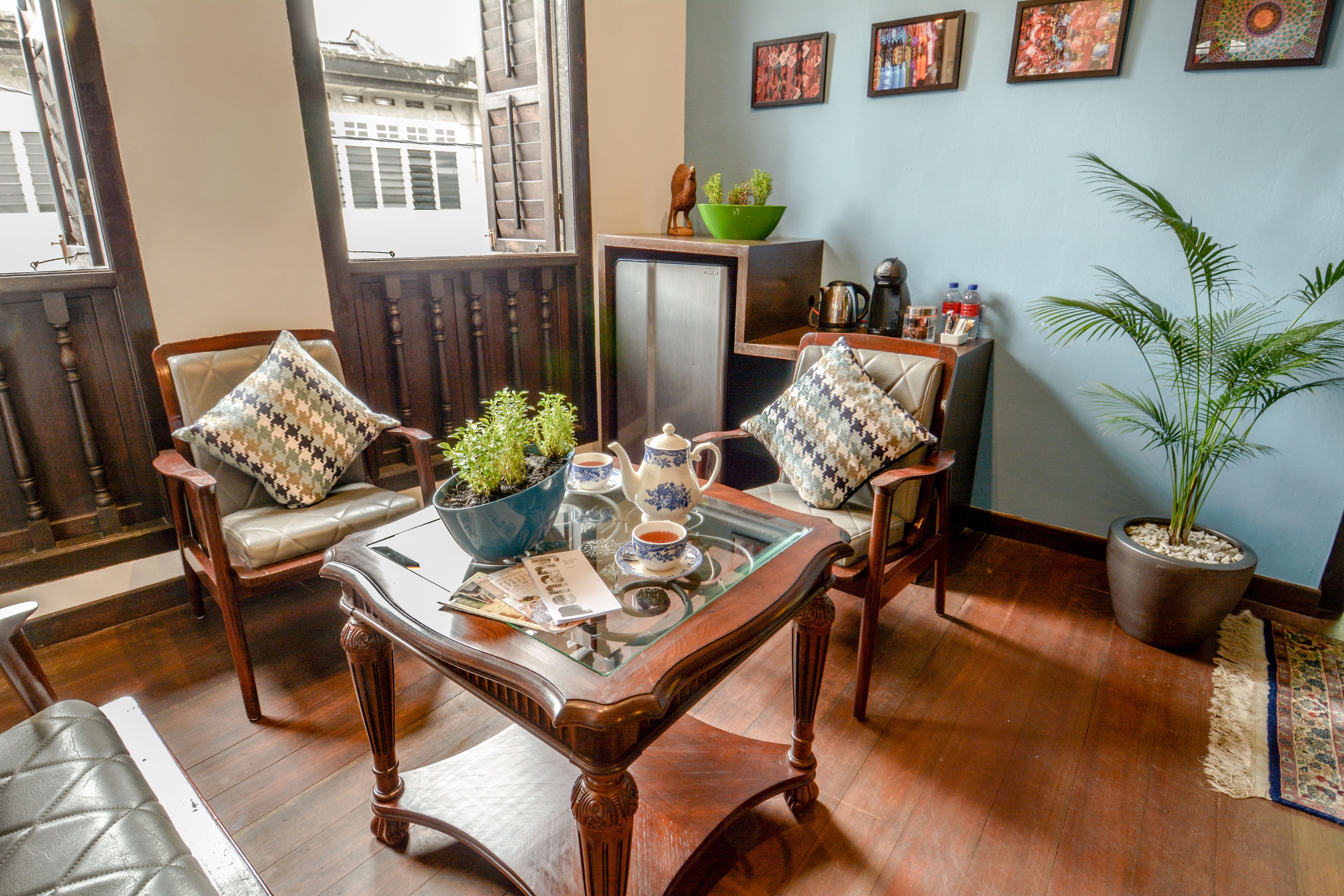 Una habitación con una mesa de madera, dos sillas, un refrigerador pequeño, una planta en maceta y una ventana con vistas a edificios.