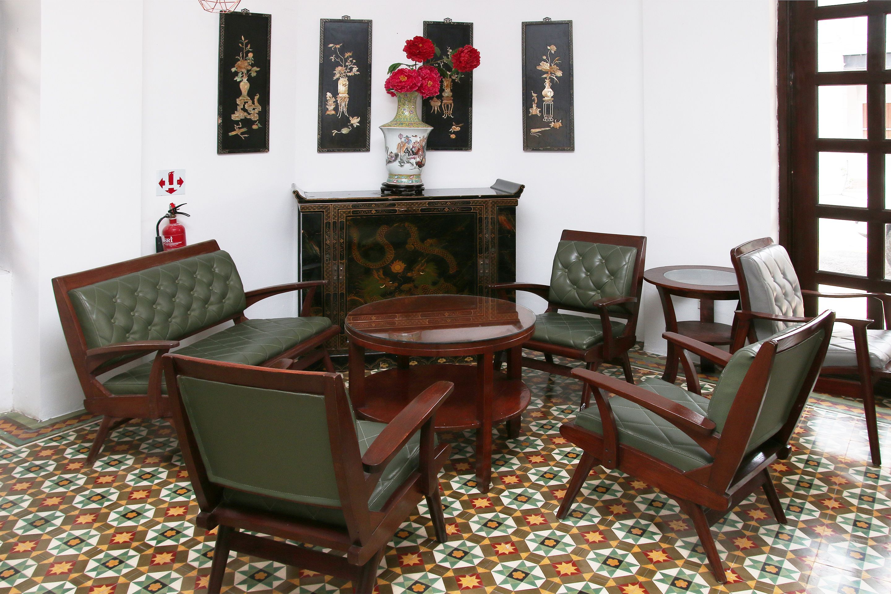 A room with a round wooden table surrounded by six green upholstered chairs, a decorative vase with red flowers on a wooden cabinet, and a patterned floor.