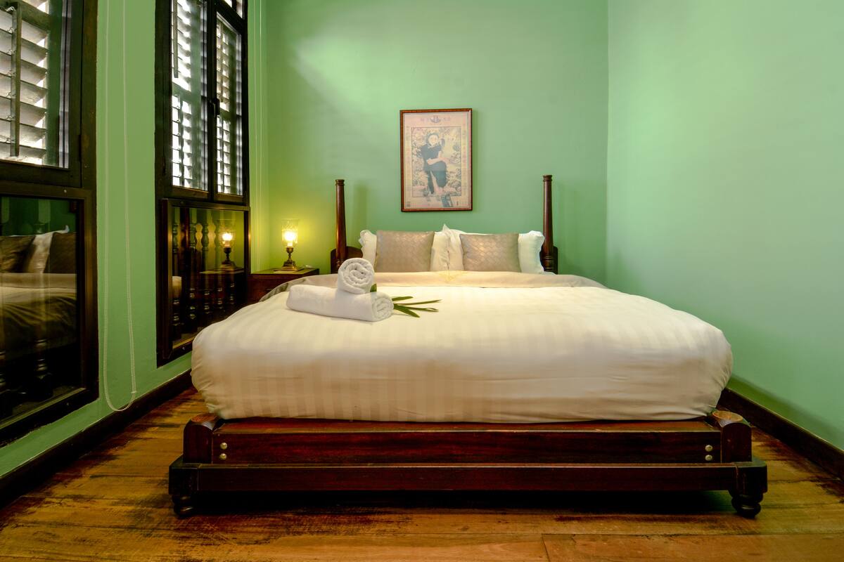 A neatly made bed with white linens and a rolled towel, a framed picture on the wall, and a wooden nightstand with a lamp.