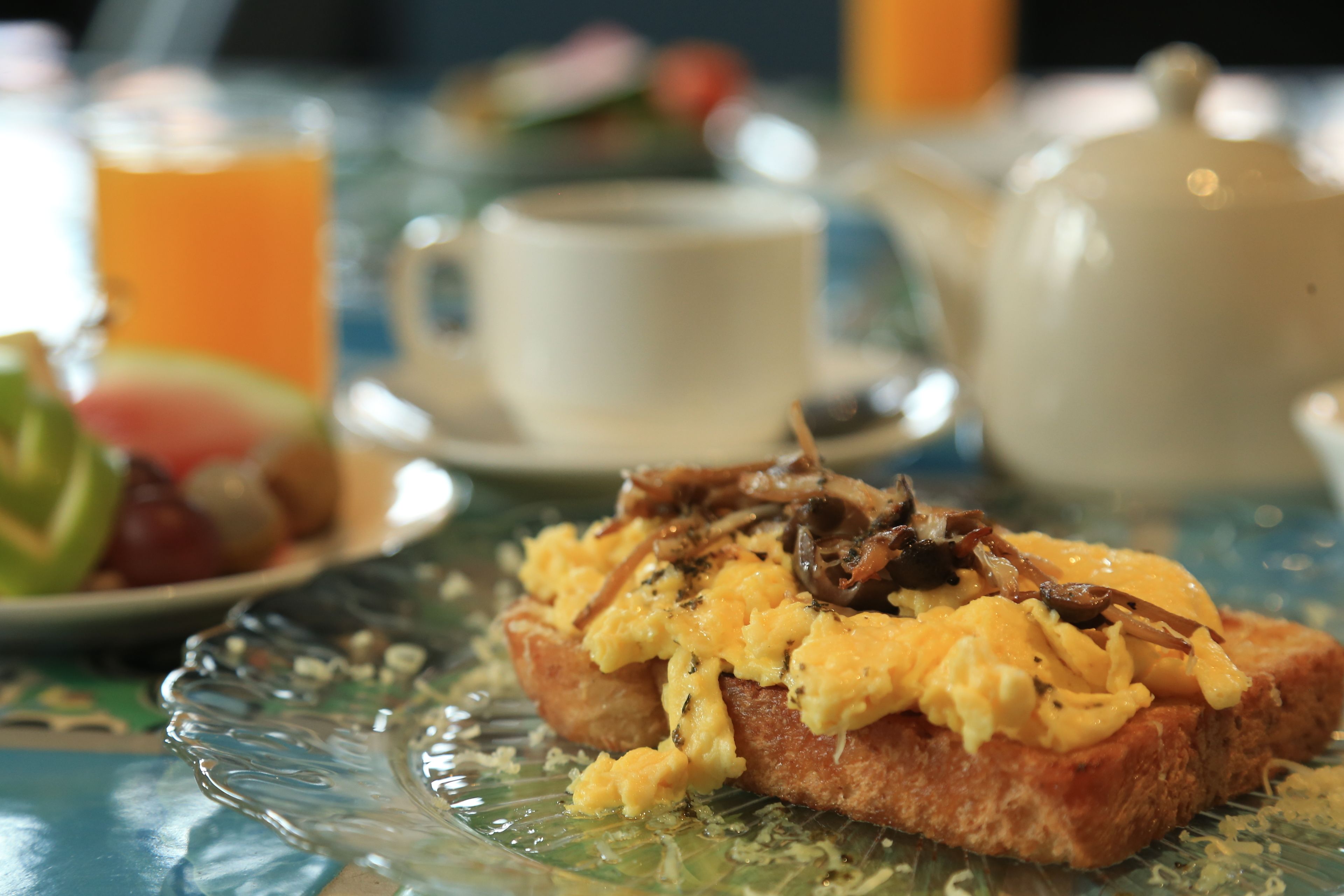 A breakfast setting with toast topped with scrambled eggs and mushrooms, a teapot, a cup, and a glass of orange juice on a table.