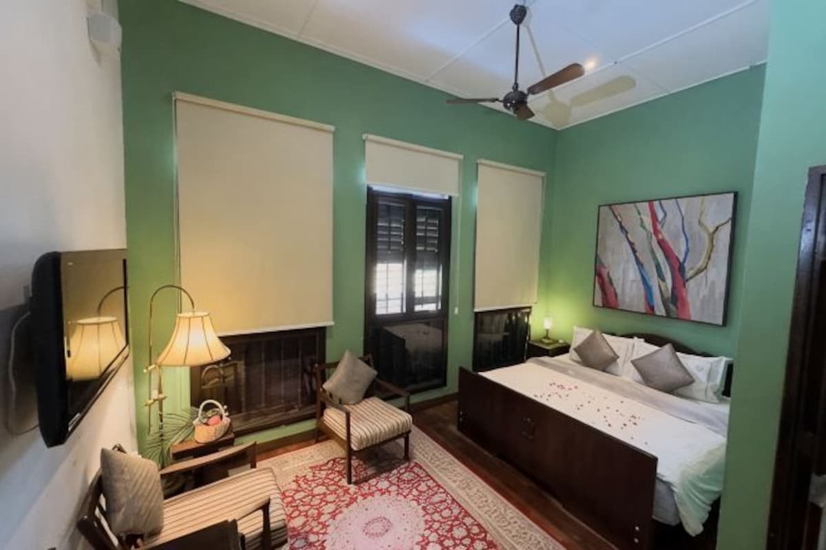 A bedroom with a bed, a chair, a lamp, a fireplace, and a painting.