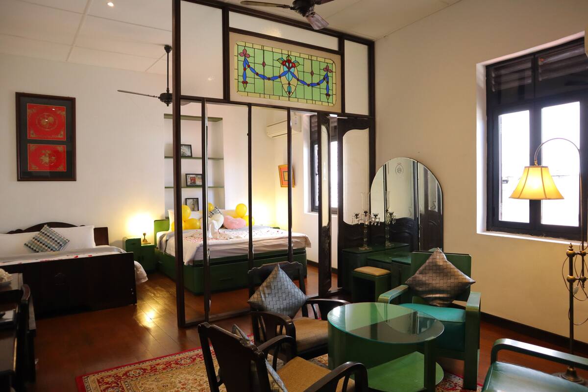 A hotel room with a bed, a desk, a chair, a glass table, a mirror, and a window with a lamp.