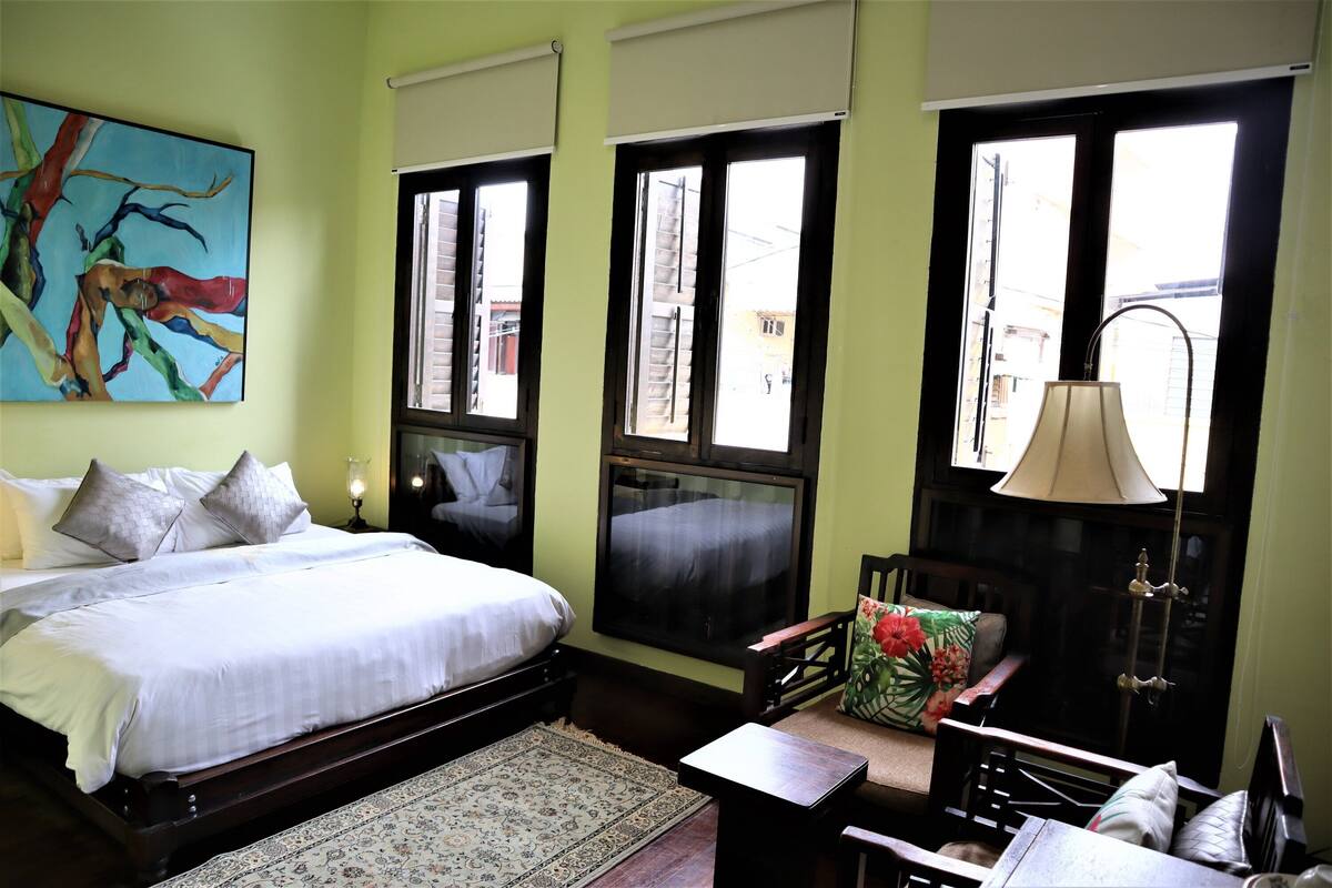 A bedroom with a bed, a desk, a chair, a lamp, and a painting on the wall.