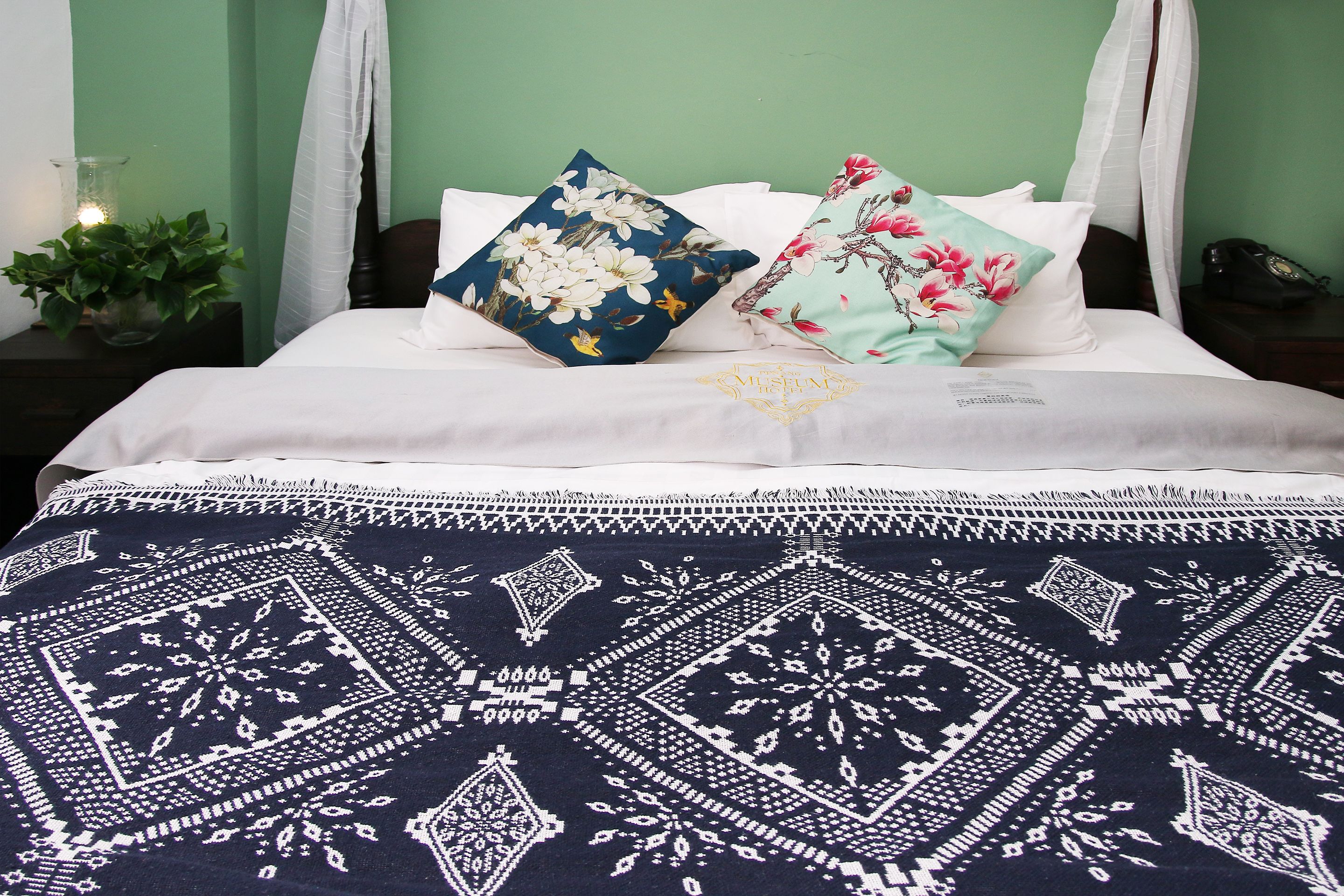 A neatly made bed with white bedding and decorative pillows, a patterned throw blanket, and a bedside table with a lamp and a plant.