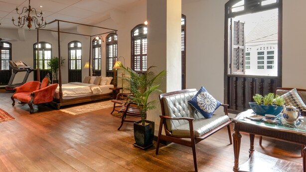 A spacious room with a large bed, a sitting area with a chair and table, a treadmill, and large windows with a view of buildings outside. - Museum Hotel (George Town)
