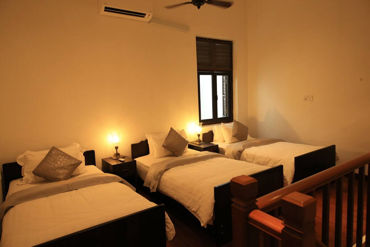 A hotel room with three single beds, each with a nightstand and a lamp. There is a window with blinds, an air conditioning unit on the wall, and a wooden railing in the foreground.