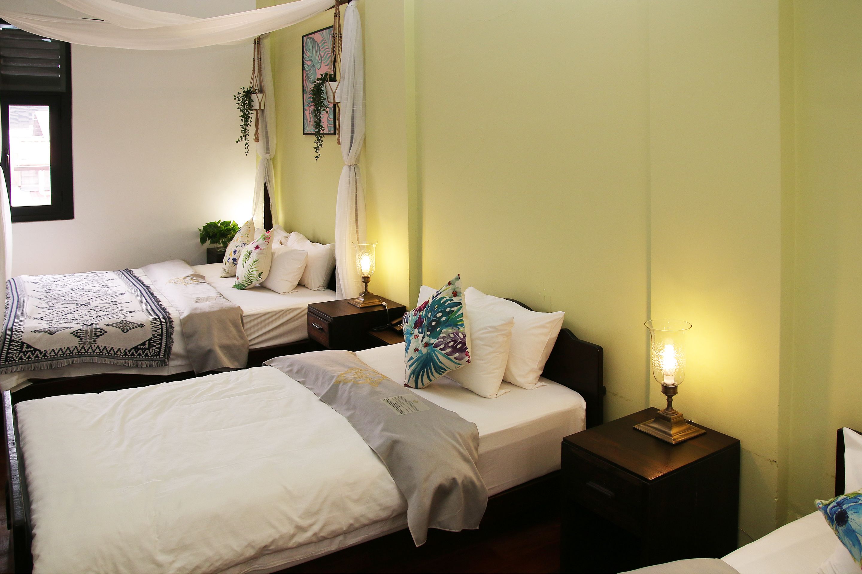 Una habitación de hotel con dos camas, cada una con ropa blanca y almohadillas decorativas. Hay mesitas de noche con lámparas y una pequeña planta sobre una de ellas.