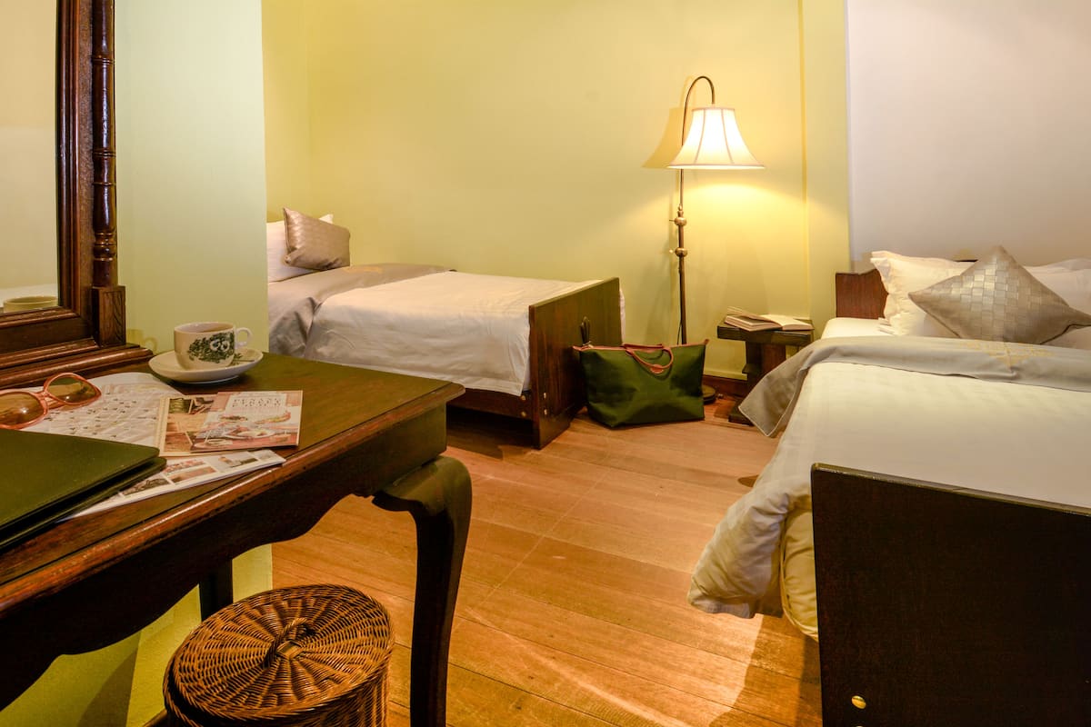 A hotel room with two single beds, a wooden desk with a chair, a lamp, and a wicker basket.