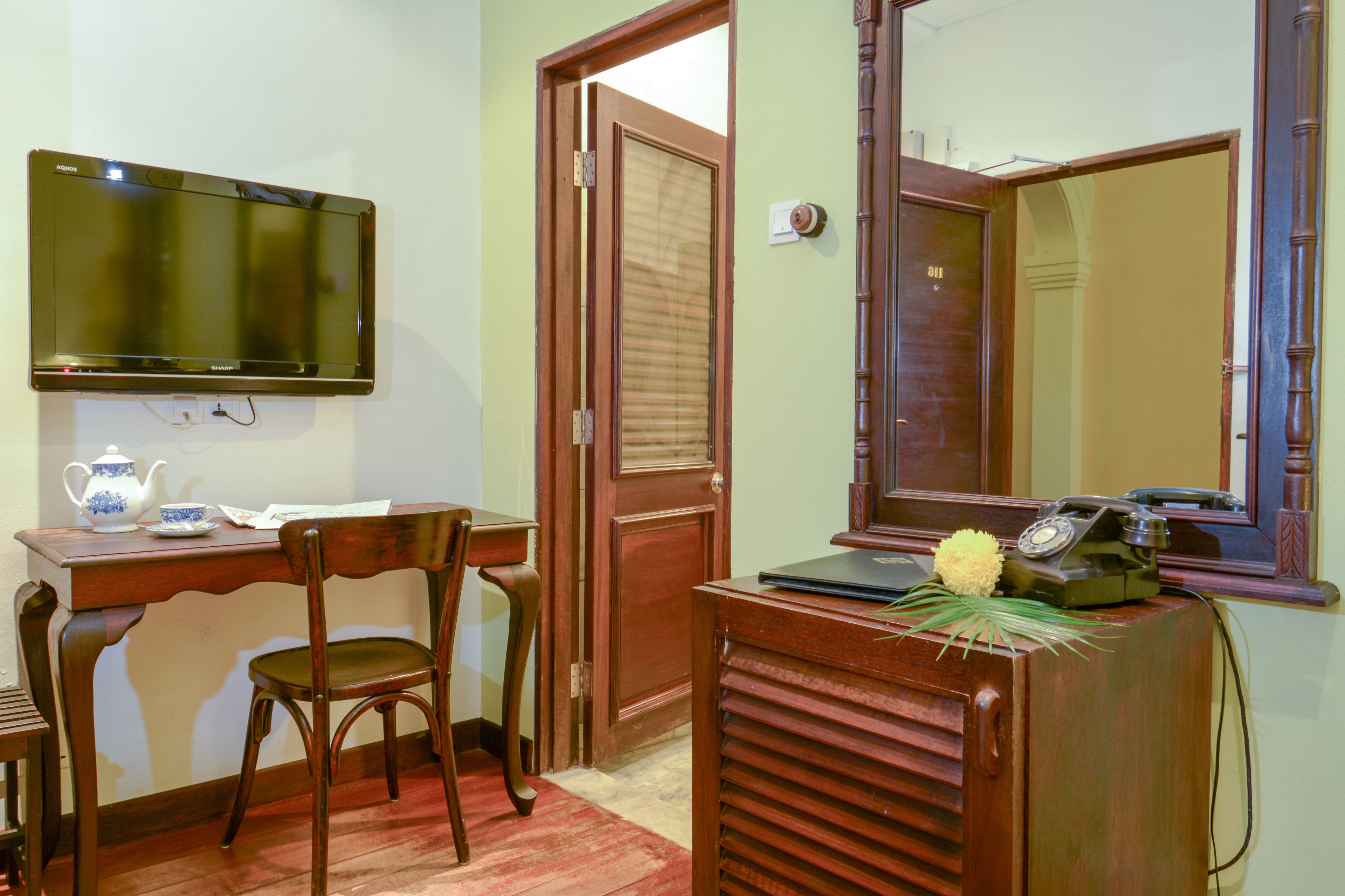 A room with a wooden desk, a chair, a television mounted on the wall, a door, and a mirror.