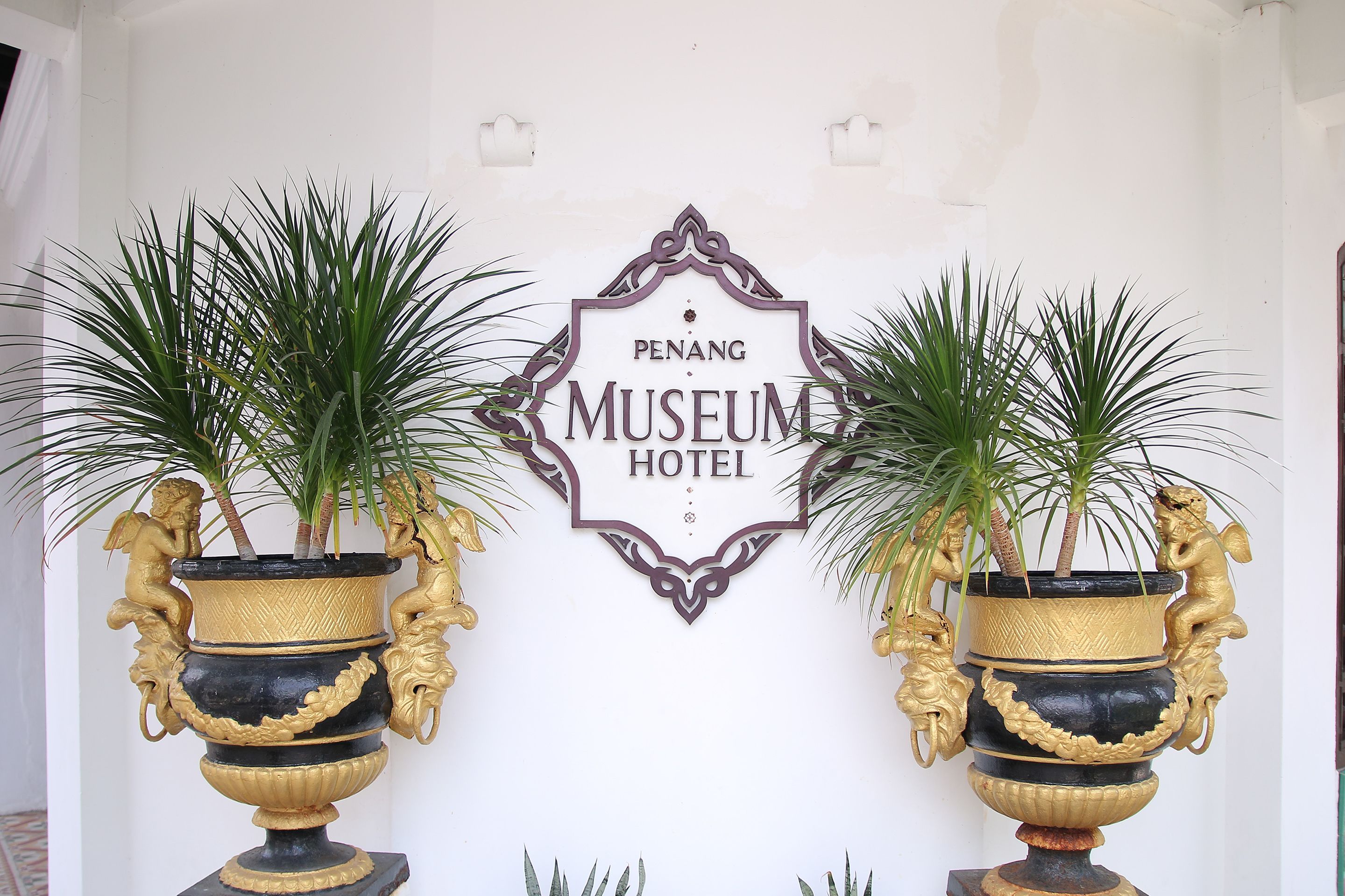 A sign for Penang Museum Hotel with two ornate vases with plants.