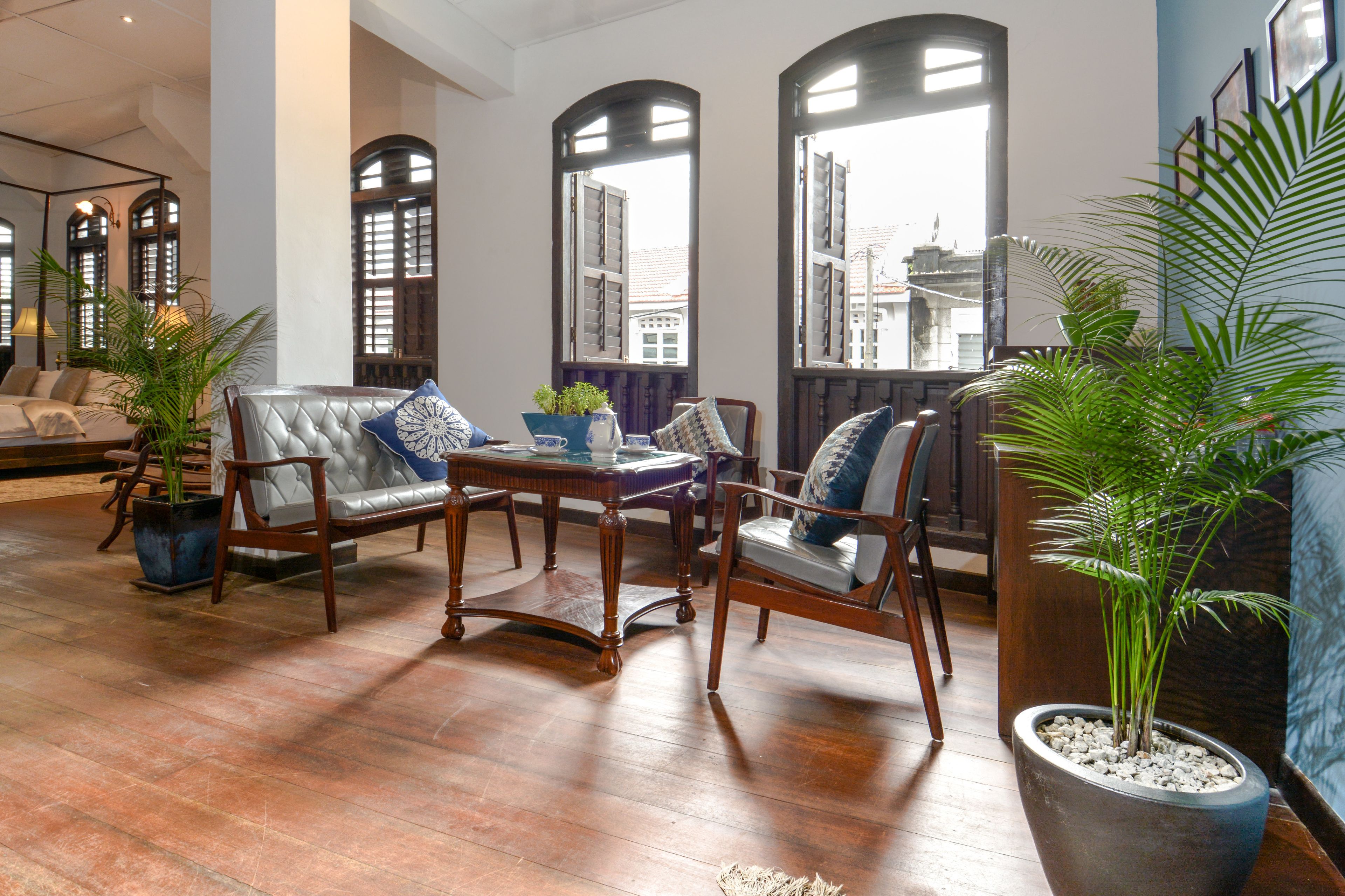 A spacious living area with a wooden floor, leather sofas, and armchairs, surrounded by large windows with shutters.