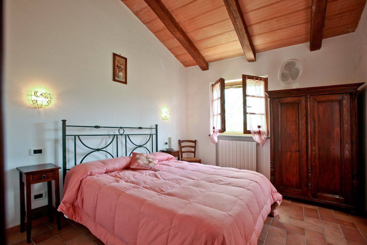 A bedroom with a bed, a wooden wardrobe, a nightstand, a wall-mounted light, and a window with curtains.