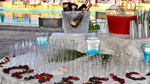 Mesa dispuesta con vasos, un balde de hielo y una bebida roja, con una piscina y sombrillas al fondo.